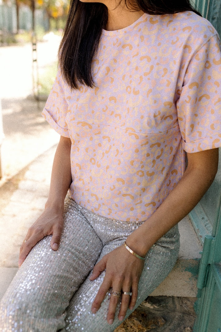 Person wearing a light pink t-shirt with a subtle leopard pattern and silver pants sitting outdoors.