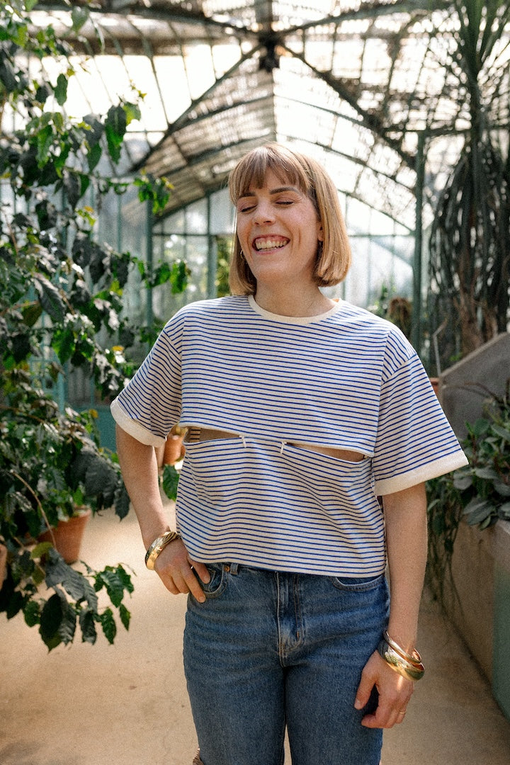 Woman standing in a greenhouse wearing a pair of jeans and a striped t-shirt with openings for nursing.
