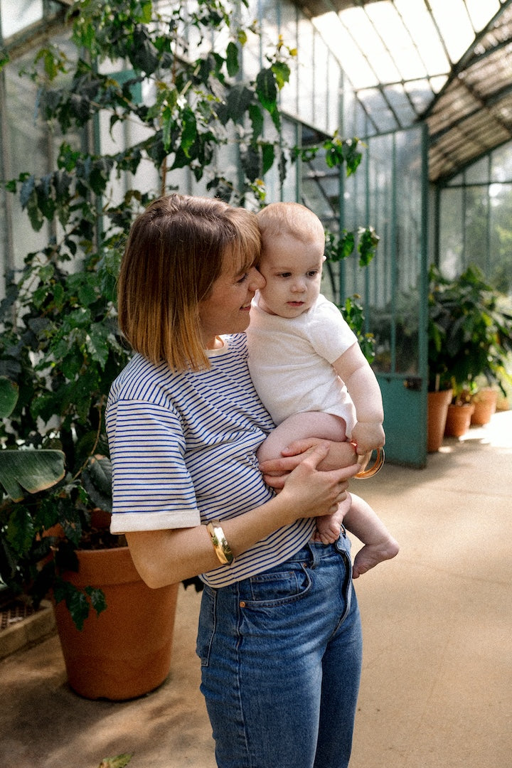 Woman holding a baby in a greenhouse filled with plants with blue and white striped top