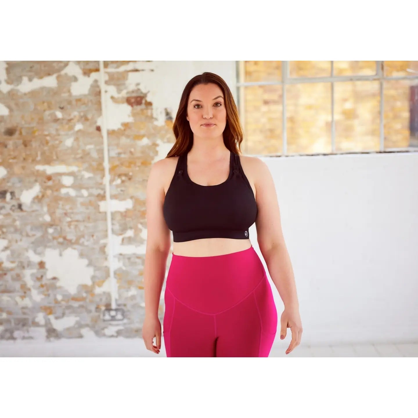 Woman wearing a black sports bra and pink leggings standing in front of a brick wall.