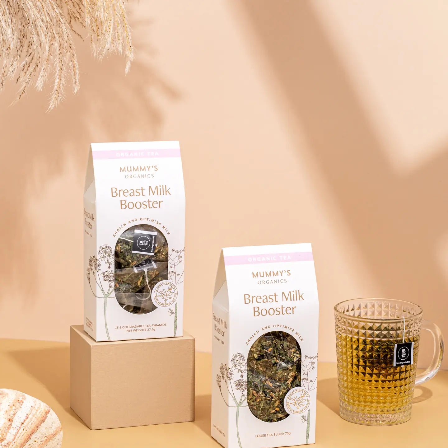 Packaging of 'Mummy's Breast Milk Booster' tea with loose tea visible through a clear window, displayed next to a glass of tea. The boxes are predominantly white with floral patterns and text detailing the product name and benefits.