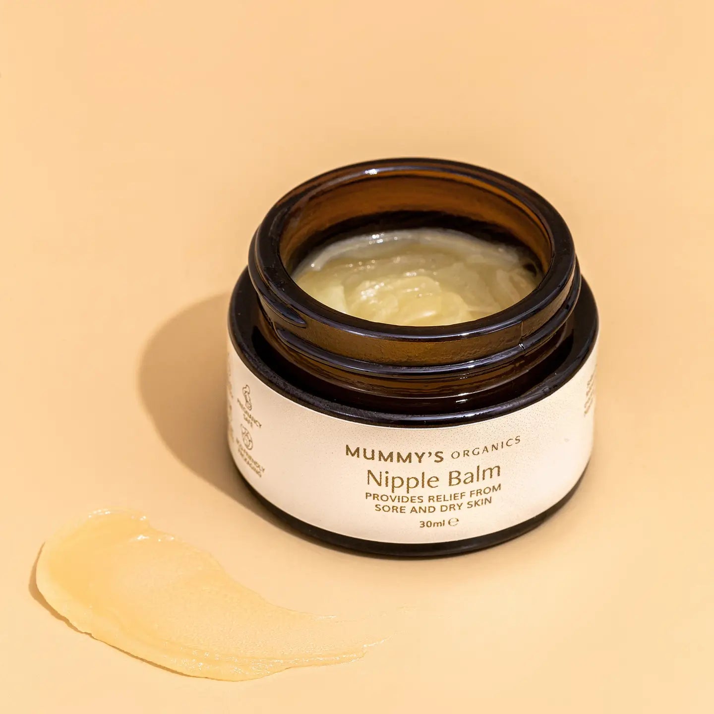 An open jar of Mummy's Organics Nipple Balm with the balm spread out in front of the jar on a beige background.