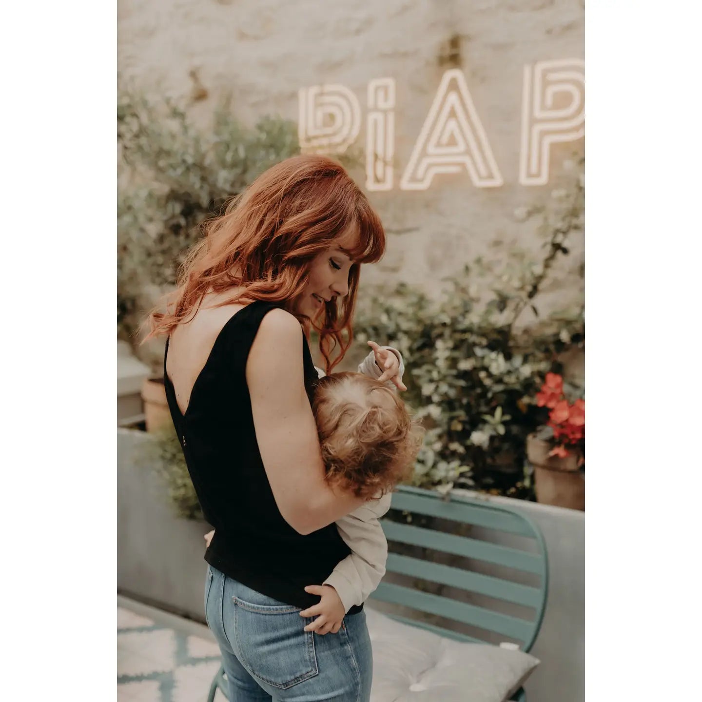Woman with black top holding a child outdoors with plants and a bench in the background