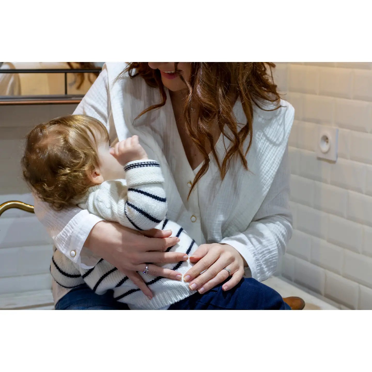 Woman wearing a white blouse holding a child in a striped sweater in a bathroom.
