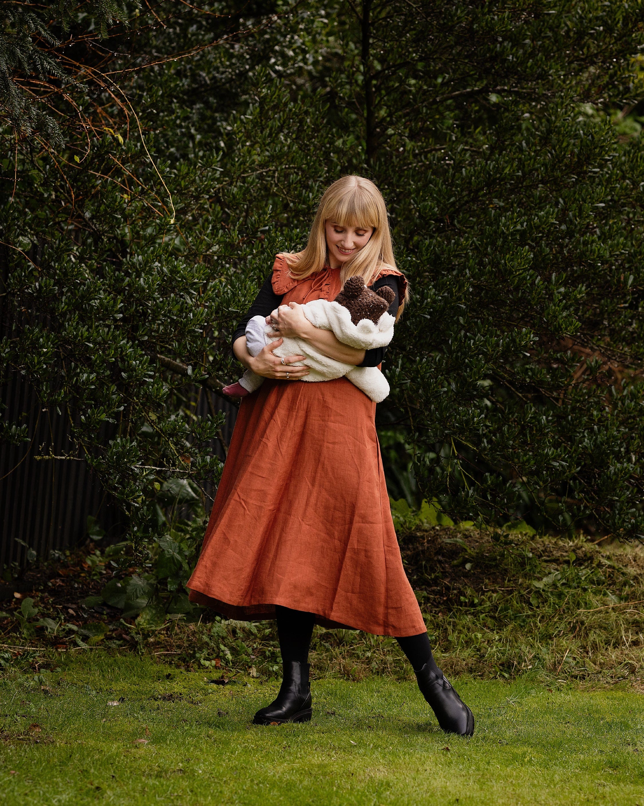 Woman in a rust-colored dress holding a baby with a teddy bear hat outdoors.