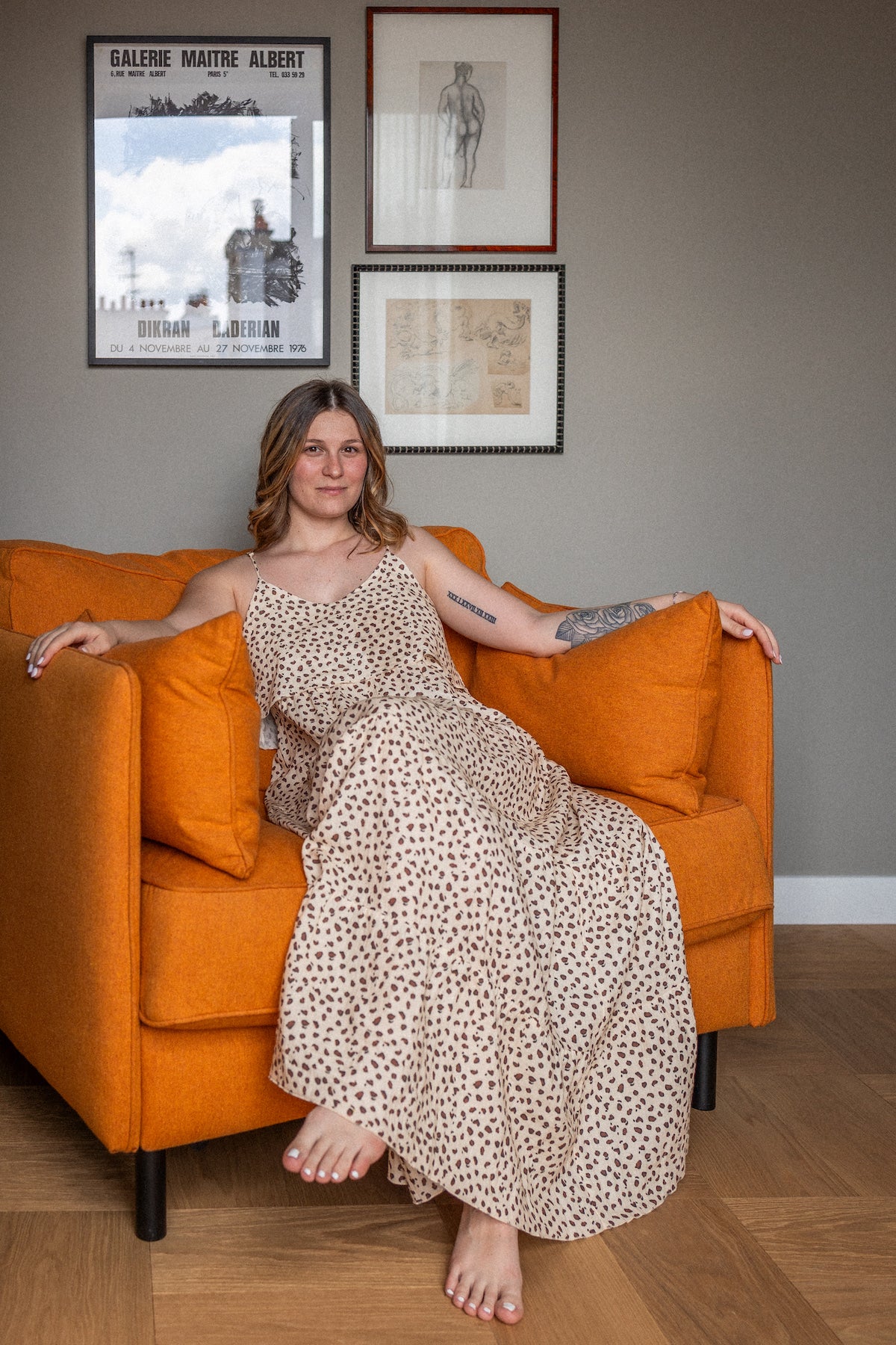 Woman in a leopard print patterned dress sitting on an orange couch with framed pictures on the wall.