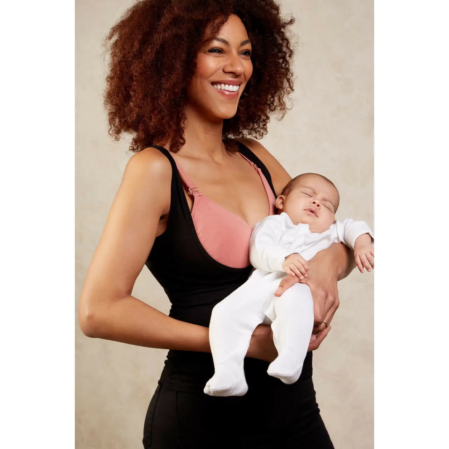 Woman holding a baby wearing a black underbust top and pink nursing bra against a neutral background