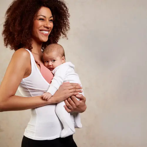 Woman holding a baby wearing a white underbust top and pink nursing bra against a neutral background