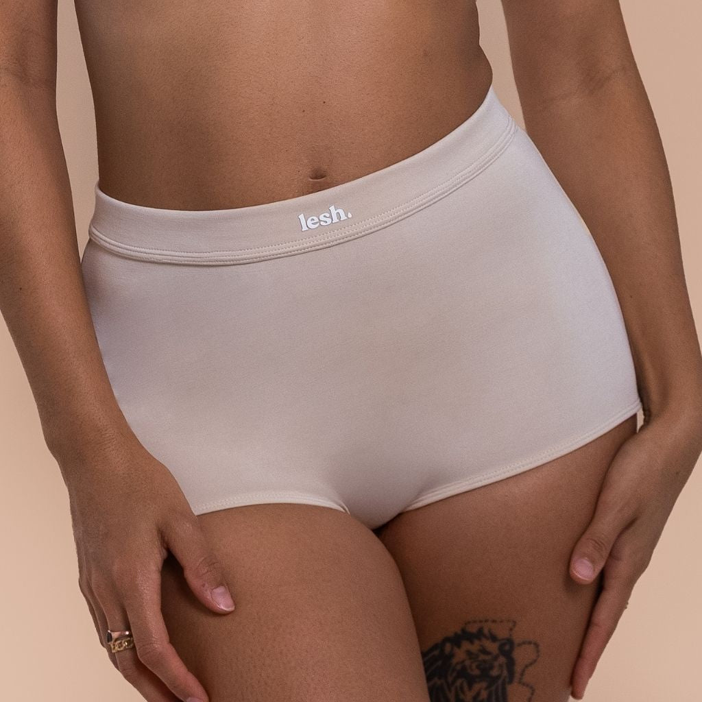 Woman wearing beige high-waisted underwear with 'lesh' branding on a beige background
