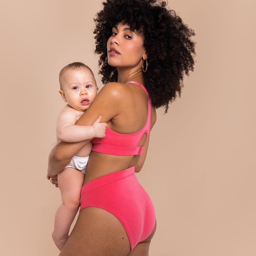 Woman in a pink underwear set holding a baby against a beige background