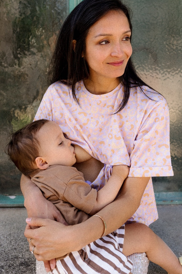 Woman breastfeeding a baby outdoors wearing a pink leopard print T-shirt against a textured wall.