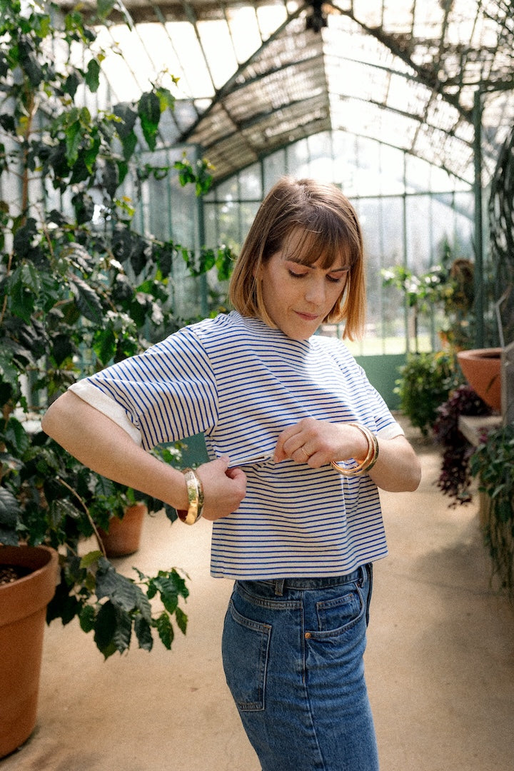 Woman in a striped T-shirt opening a nursing zip standing in a greenhouse filled with plants.