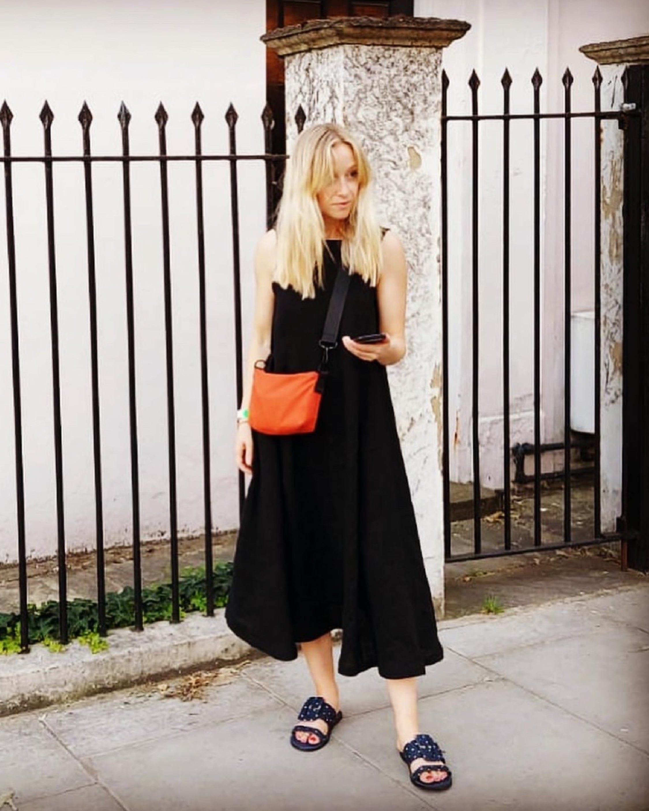 Woman in a black dress with an orange bag standing against a white wall with black iron railing.