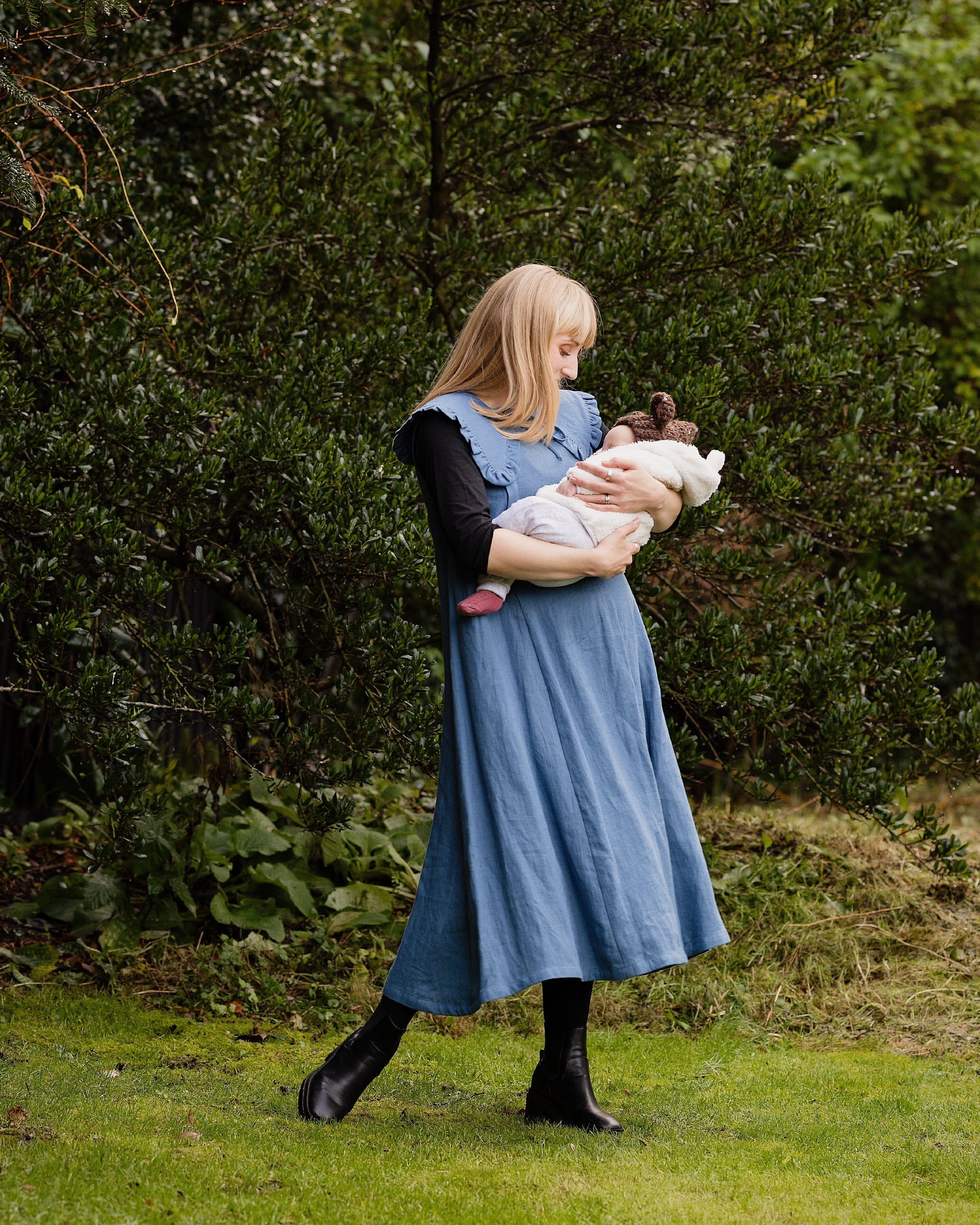 Woman in a blue dress holding a baby outdoors with greenery in the background