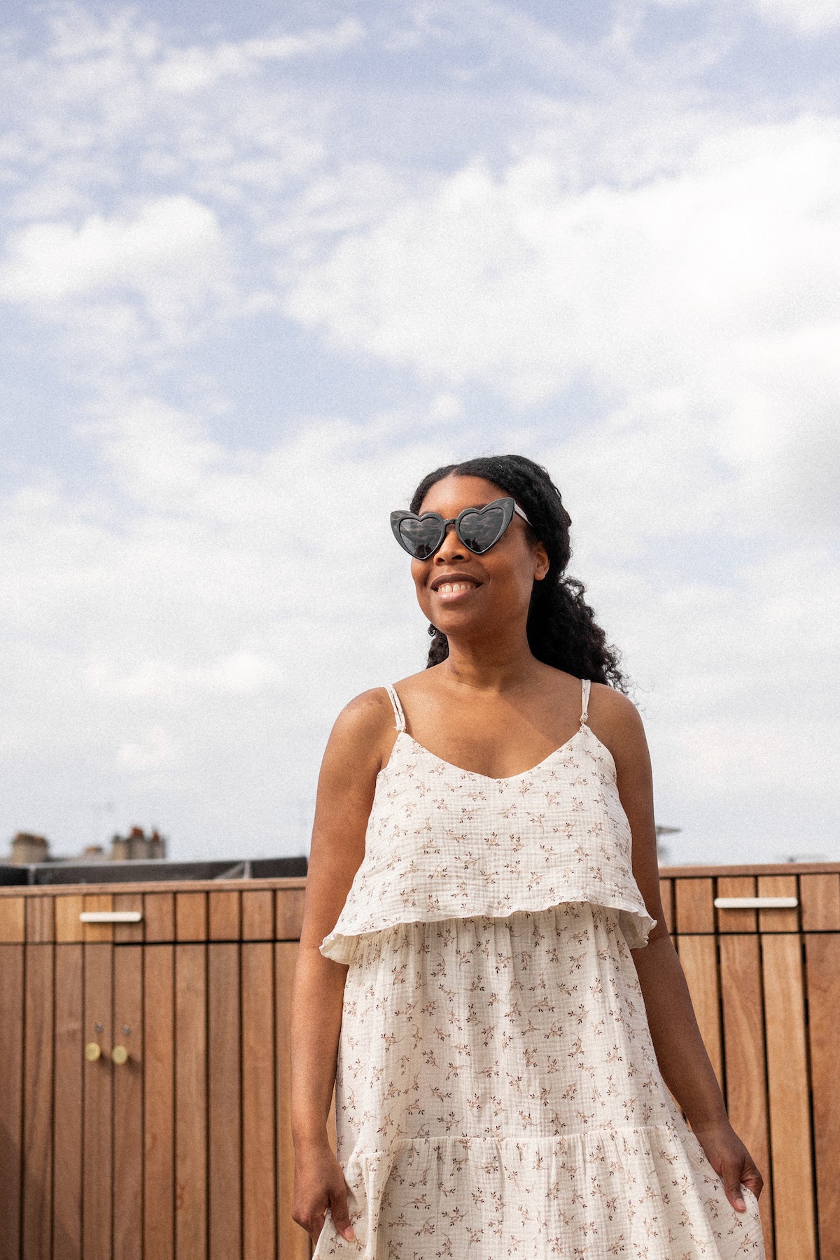 Woman in a long white and flowery dress with sunglasses standing on a rooftop with a cloudy sky.