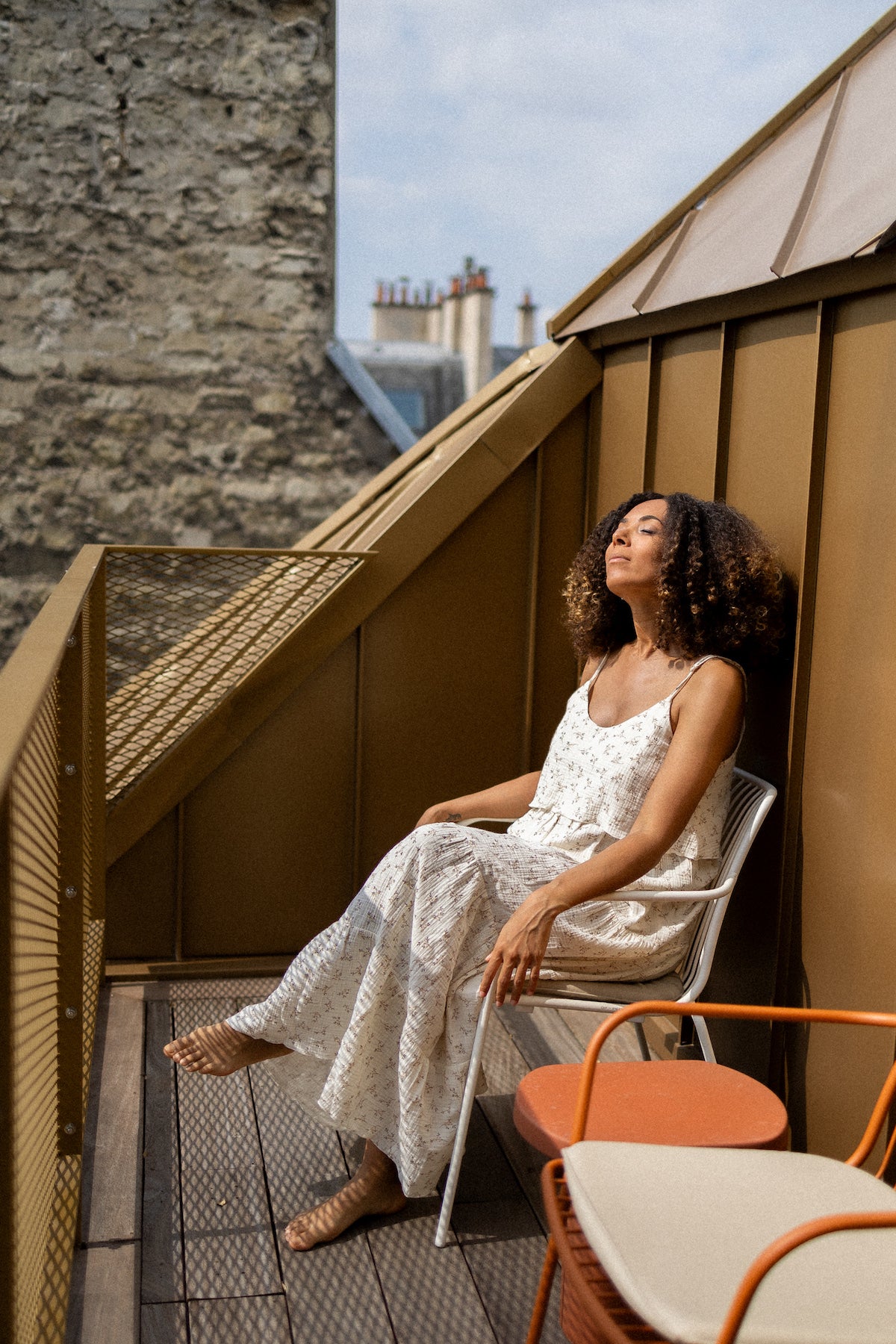 Woman in a long white and flowery dress sitting on a rooftop bench with a cloudy sky.