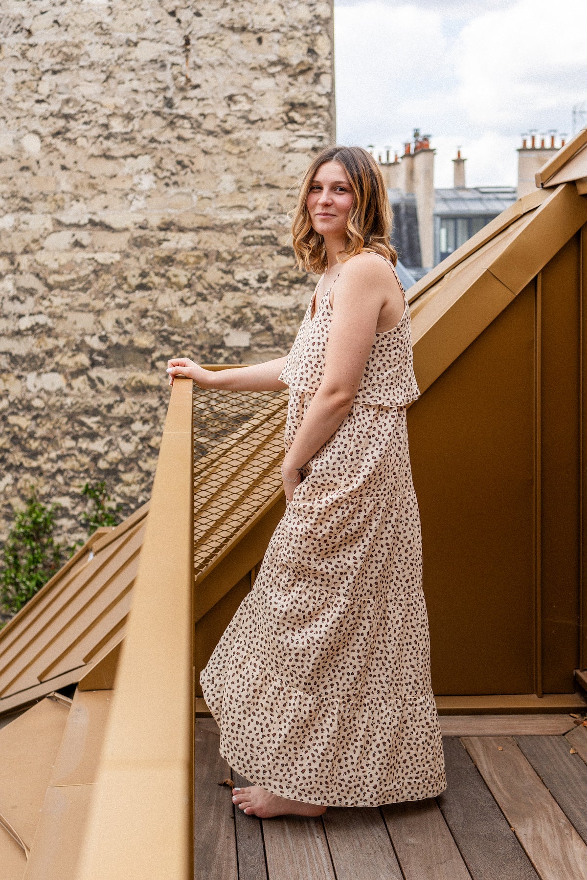 Woman in a leopard print long dress standing on a wooden deck with stone wall and buildings in the background