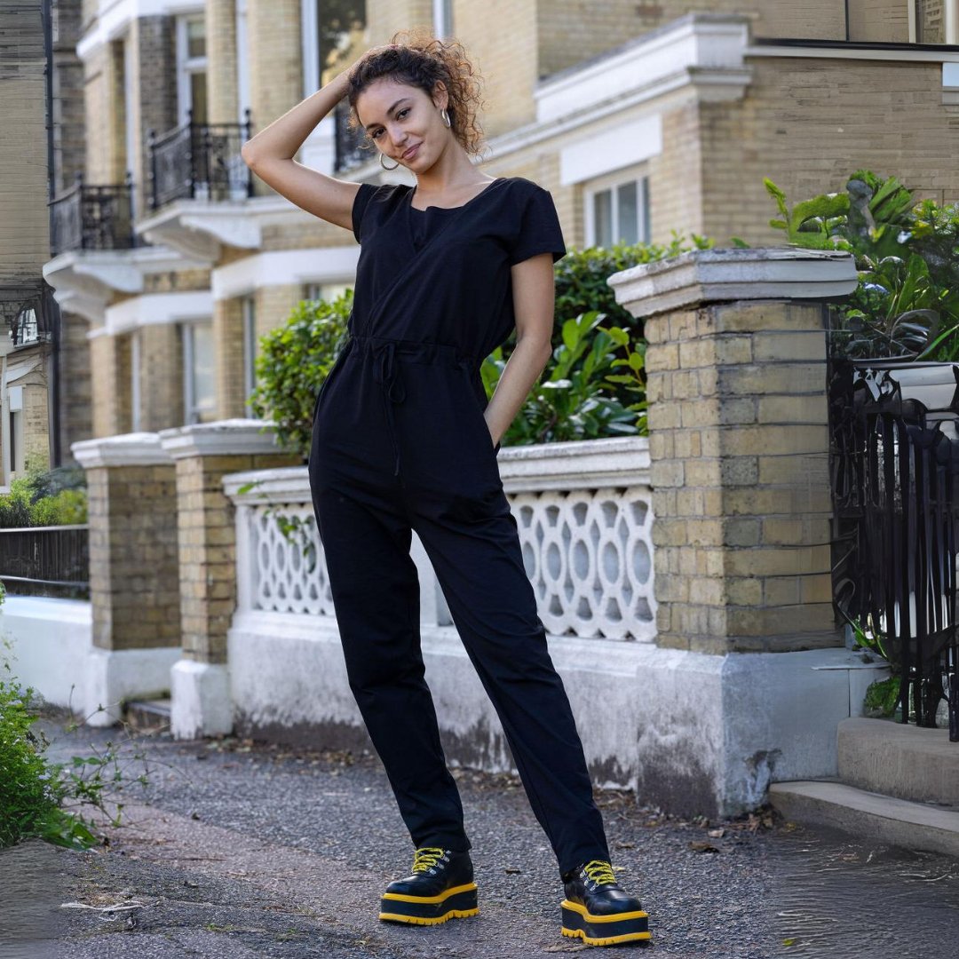 Woman in a black jumpsuit with black and yellow shoes standing outdoors.