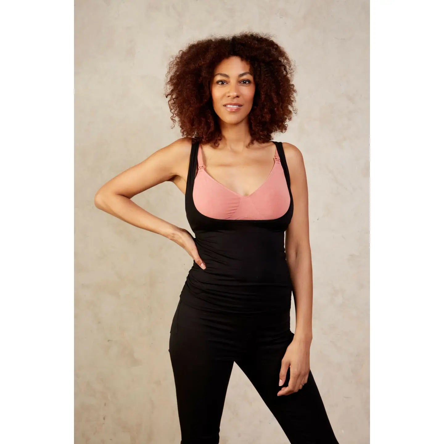 Woman wearing a black underbust top over a pink bralette with black pants against a neutral background