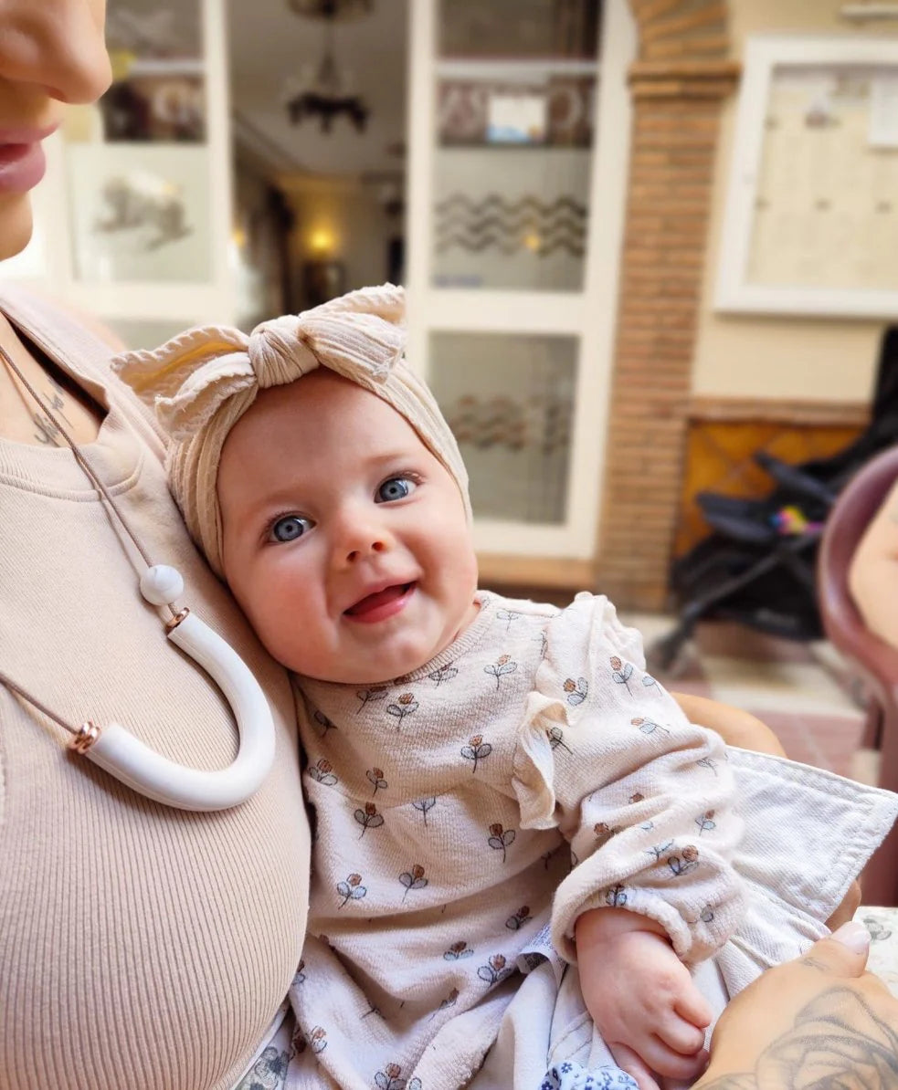 Baby in a floral outfit and headband being held by an adult wearing a breastfeeding necklace